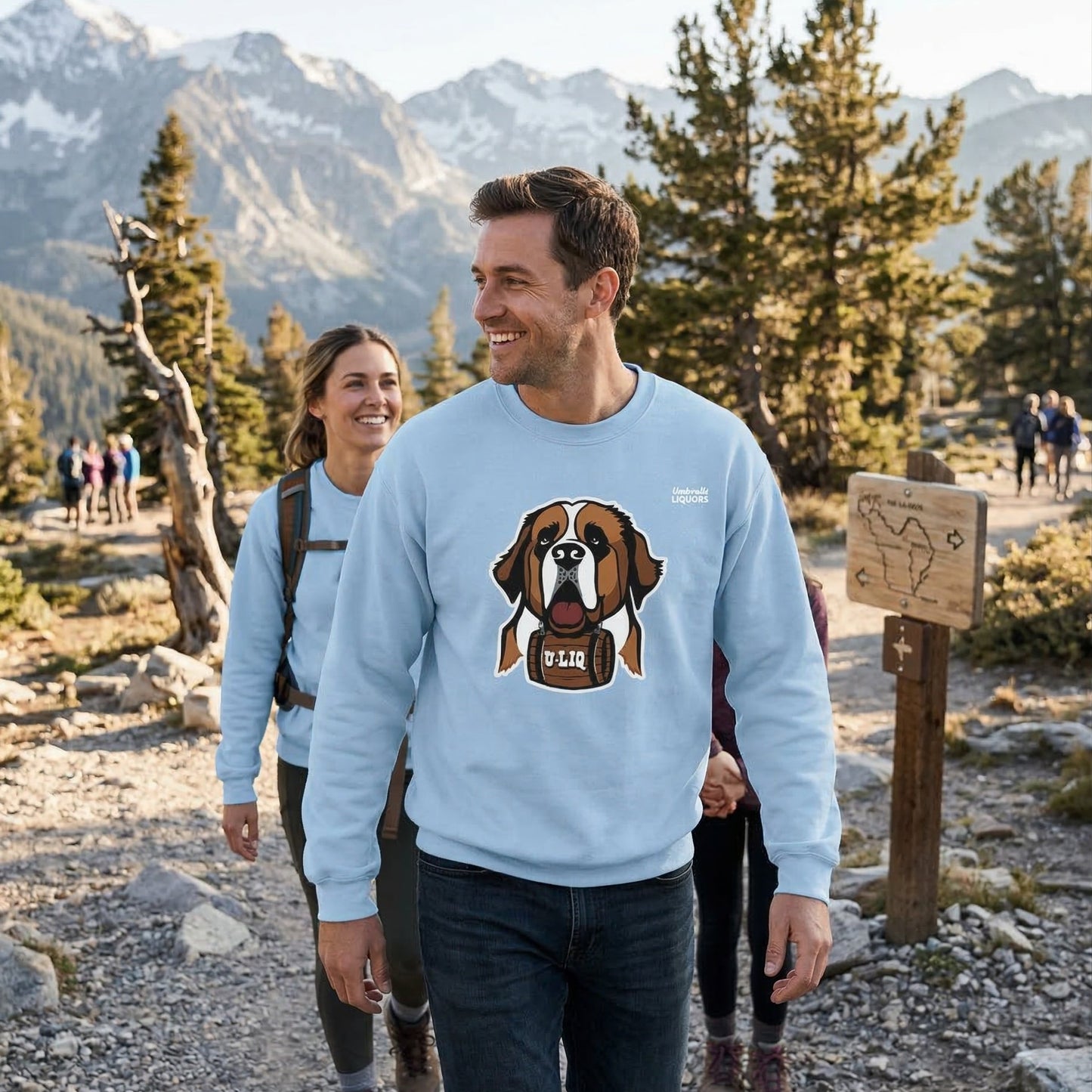 Man wearing a light blue University Liquors sweatshirt, walking on a trail with mountains in the background.