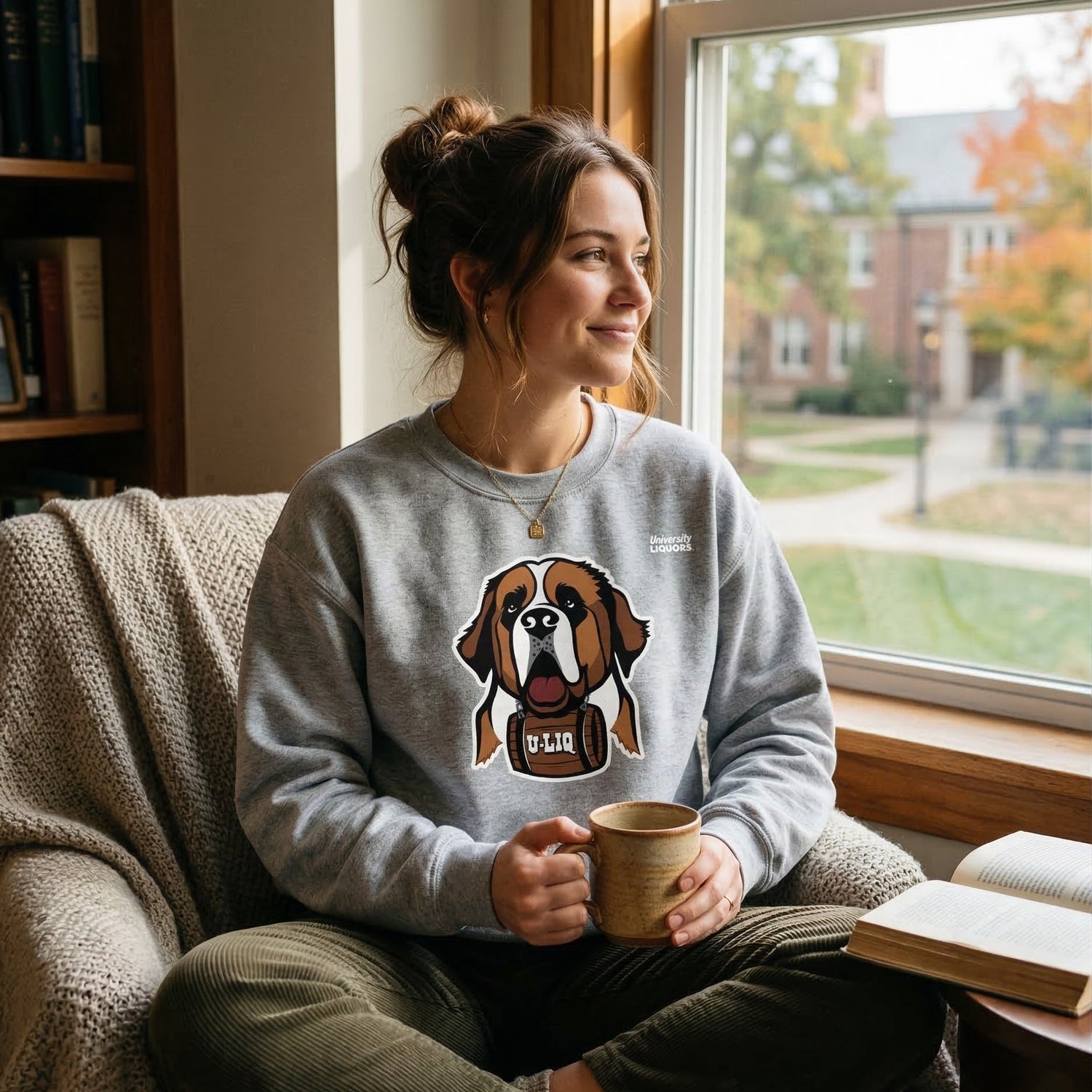 Woman sitting on a couch holding a mug, wearing a gray University Liquors sweatshirt, in a cozy room with a window view.