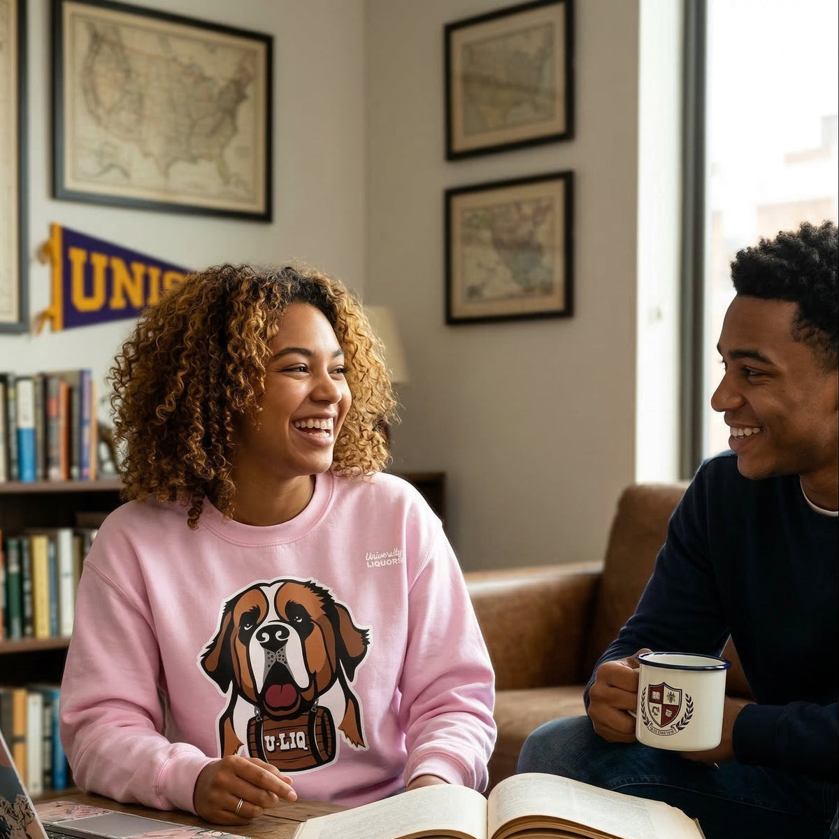 Two people sitting at a table wearing light pink University Liquors sweatshirts with books and a laptop, smiling and laughing.