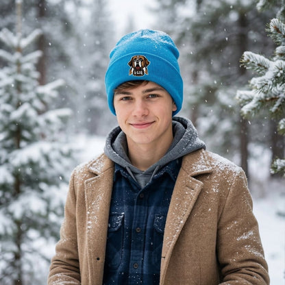 Person wearing a blue beanie with a University Liquors logo, brown coat, and dark shirt in a snowy forest.
