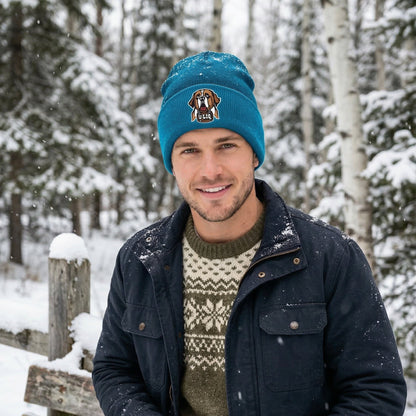 Man wearing a blue beanie with a University Liquors logo and dark jacket in a snowy forest