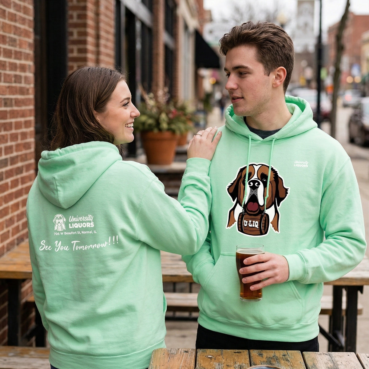 Two people wearing mint green University Liquors hoodie on an urban street.