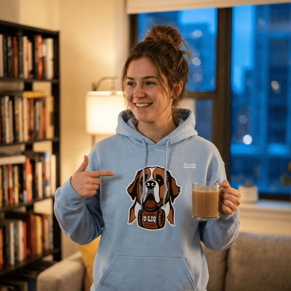 Woman wearing a light blue University Liquors hoodie, pointing to the hoodie, in a cozy living room.