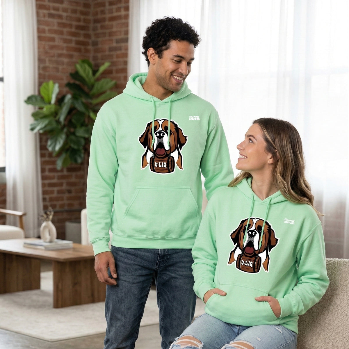 Two people wearing matching mint green University Liquors hoodies in a living room.