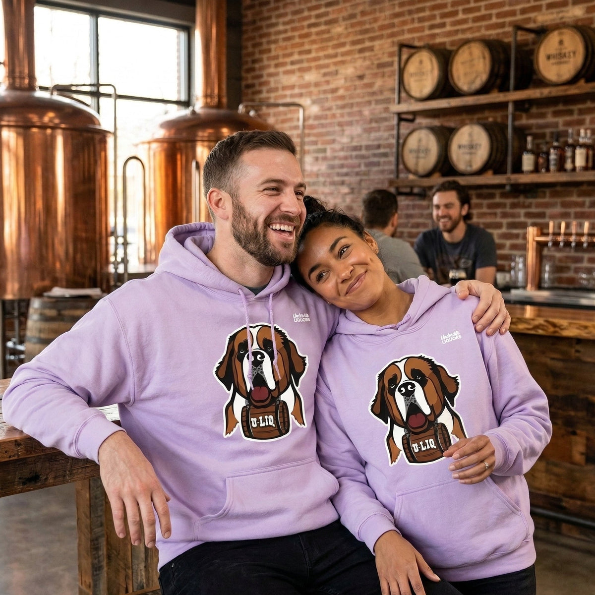 Two people wearing matching purple University Liquors hoodies in a brewery setting.